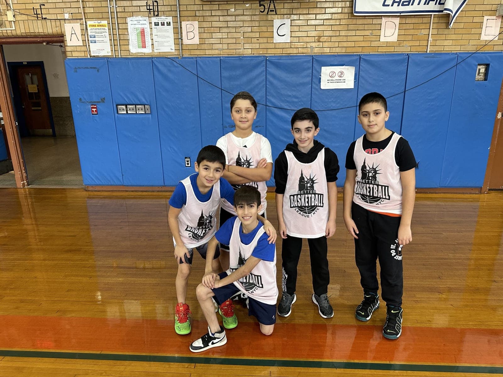 Five players posing in front of the blue padded gym wall, two kneeling in front