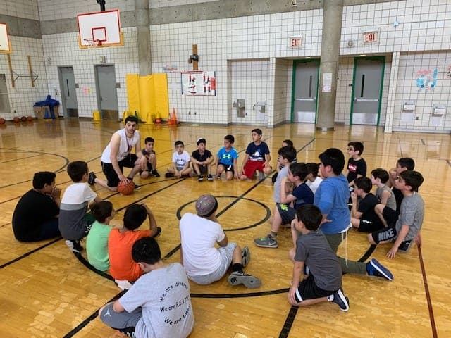 A coach kneeling with a ball, explaining a drill to players seated in a circle around him