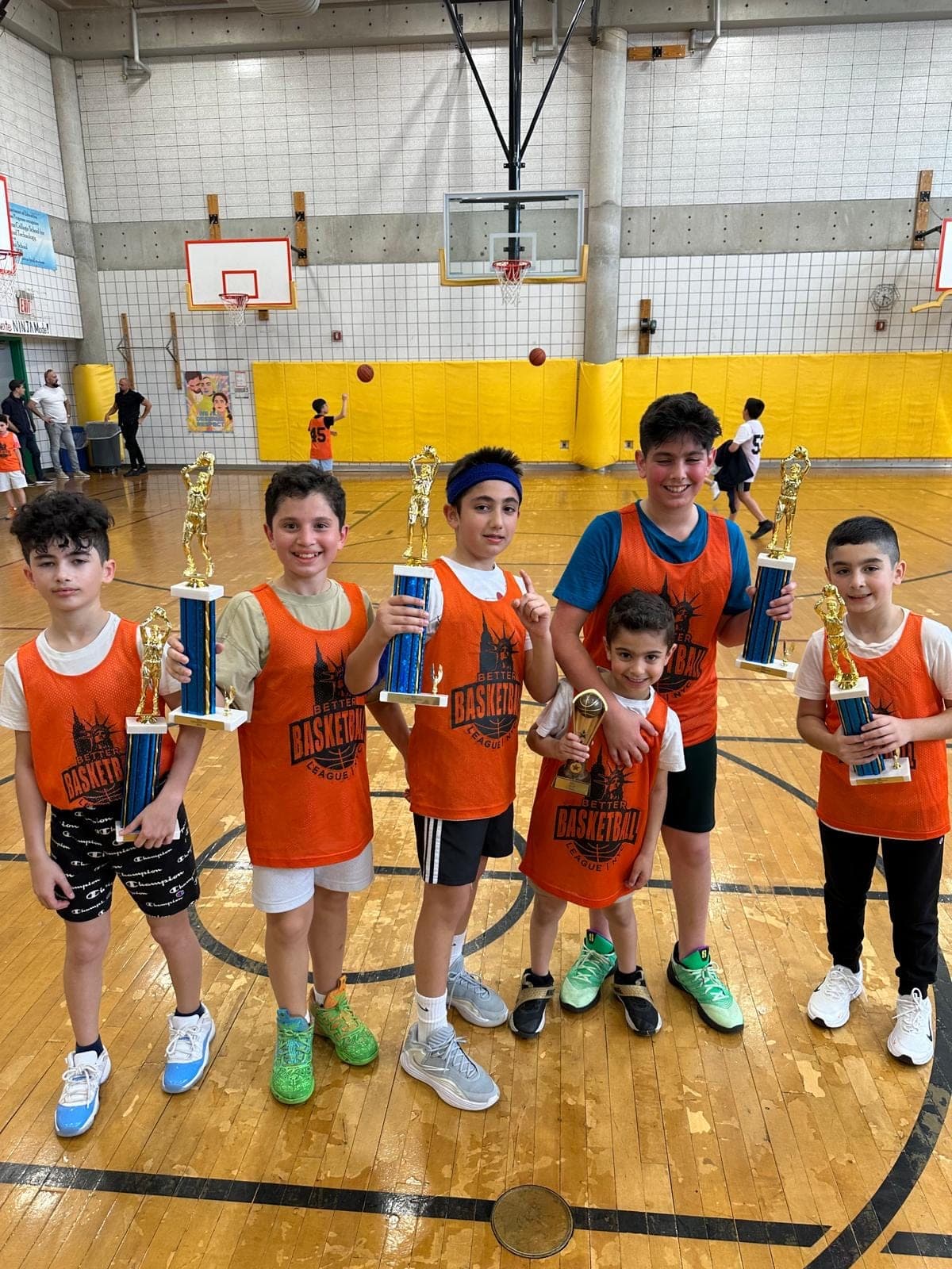 Six players in orange BBLNYC pinnies at center court, each holding a tall championship trophy
