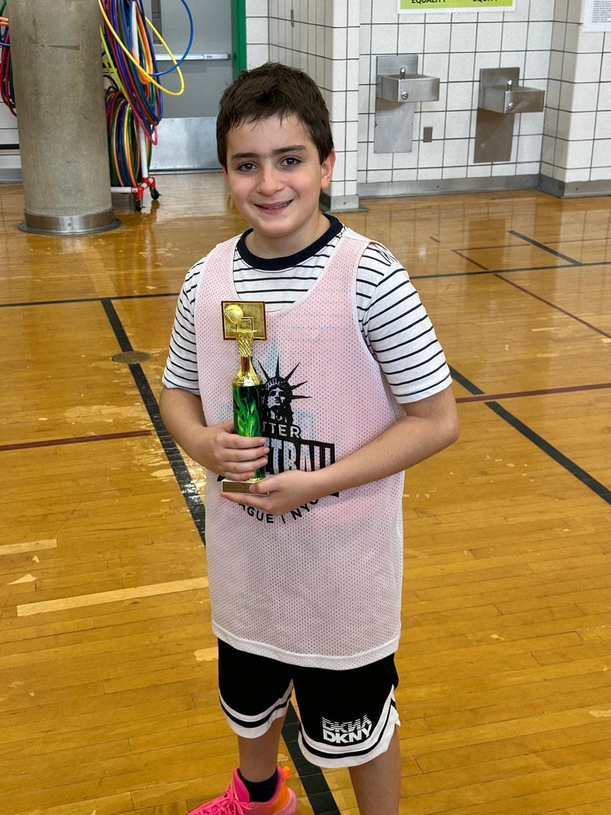 A player in a white BBLNYC pinnie holding his end-of-season trophy at the side of the gym