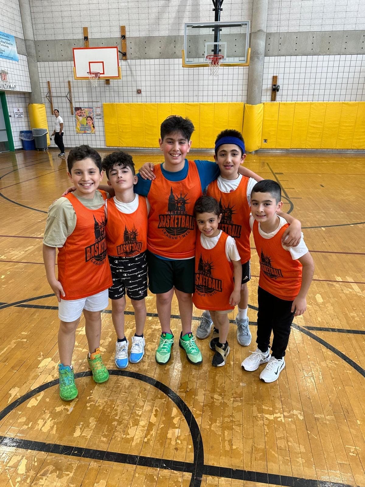 Six BBLNYC players in orange pinnies, arms around each other's shoulders, posed under the hoop after a Sunday session