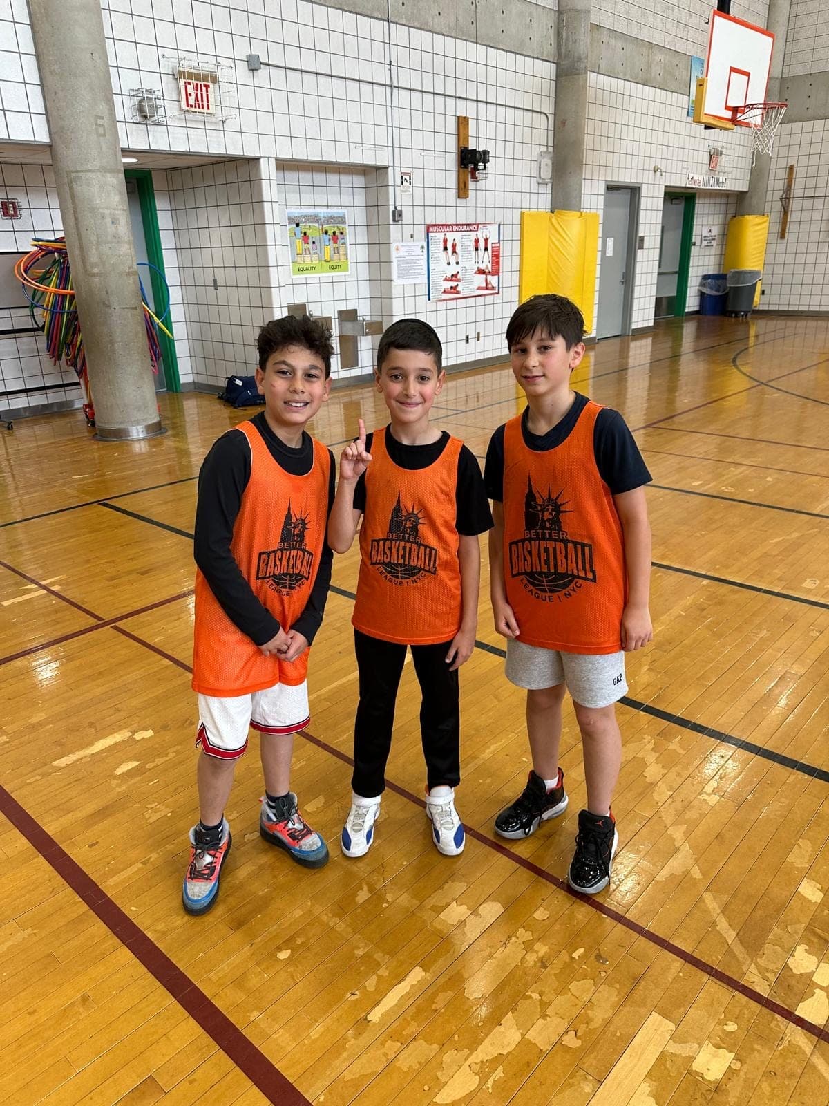 Three players in orange BBLNYC pinnies standing together at the side of the gym